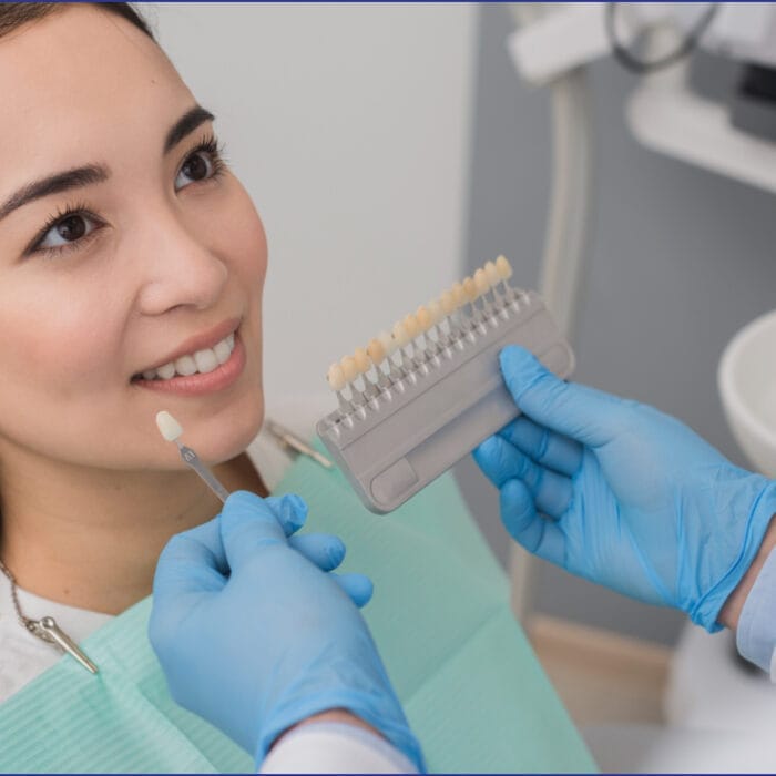 A woman sitting in a dental chair, holding a mirror and examining a set of veneer samples presented by a dentist, with a dental tray and equipment visible nearby.