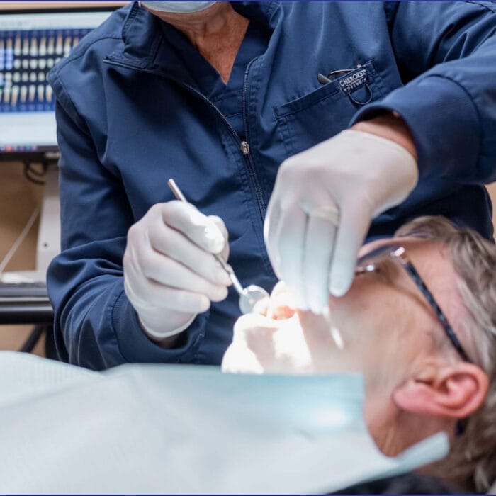 An elderly man receiving an emergency dental checkup, sitting in a dental chair while the dentist examines his teeth, with concern visible on both faces and medical equipment in the background.