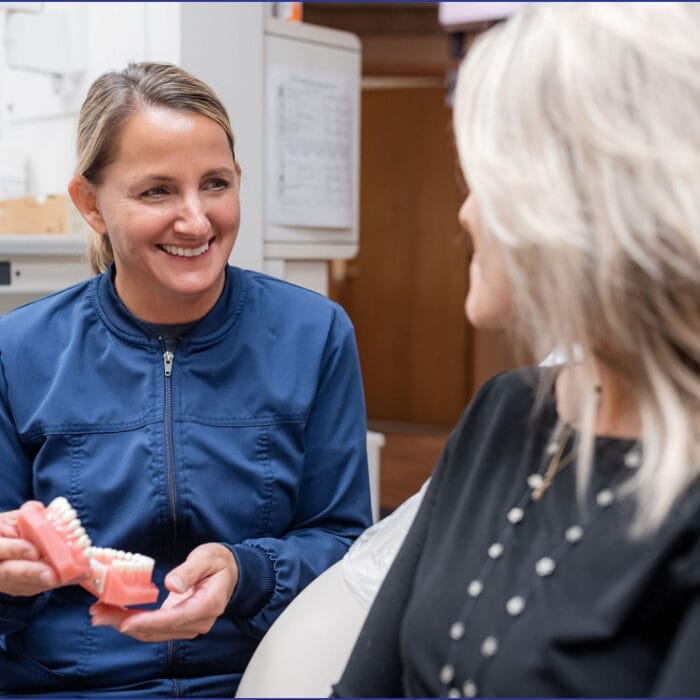 A woman dentist showing a set of dentures to an elderly woman, both seated in a dental clinic, with dental tools and equipment visible in the background.