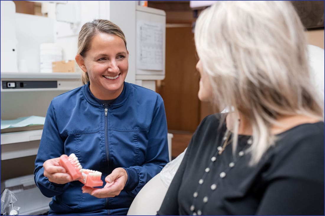 A woman dentist showing a set of dentures to an elderly woman, both seated in a dental clinic, with dental tools and equipment visible in the background.